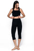 A woman modeling a black two-piece activewear set consisting of a cropped tank top and high-waisted capri leggings, standing against a plain white studio background.