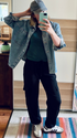 Person taking a mirror selfie wearing a denim jacket, green shirt, black pants, and white sneakers in a room.